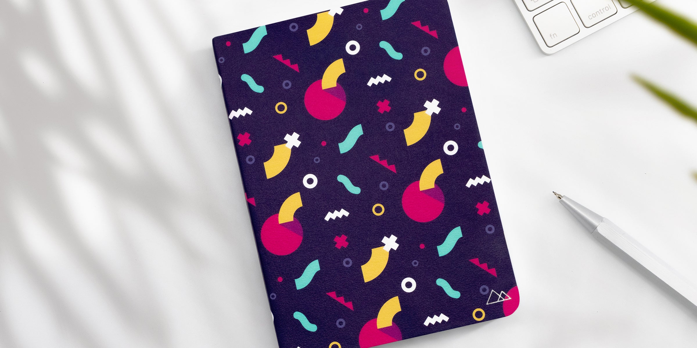 Bright and colourful notebook cover with different shapes on a white desk with a silver pen and keyboard in view.